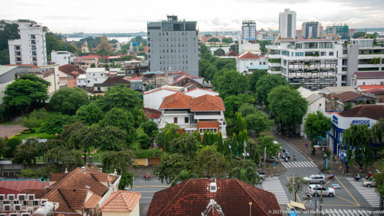 Elevated view over tree-lined streets, red-roofed villas, traffic, and mid-rise buildings in Phnom Penh.
