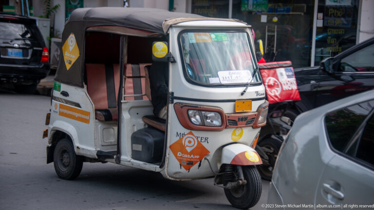 Orange-and-white motorized tuk-tuk traveling beside cars on a busy Phnom Penh street.