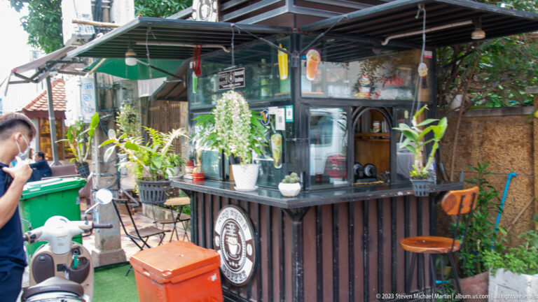 Small dark-wood coffee kiosk with plants, stools, coolers, and a parked scooter beside the street.