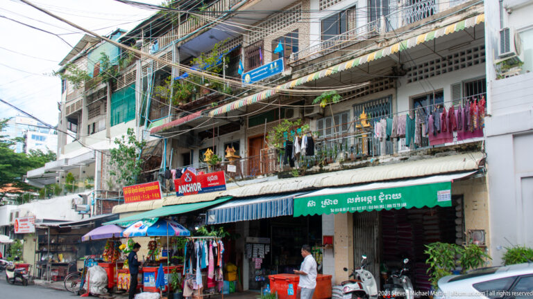 Street shops with clothing displays beneath narrow apartments, balconies, awnings, and dense overhead wiring.