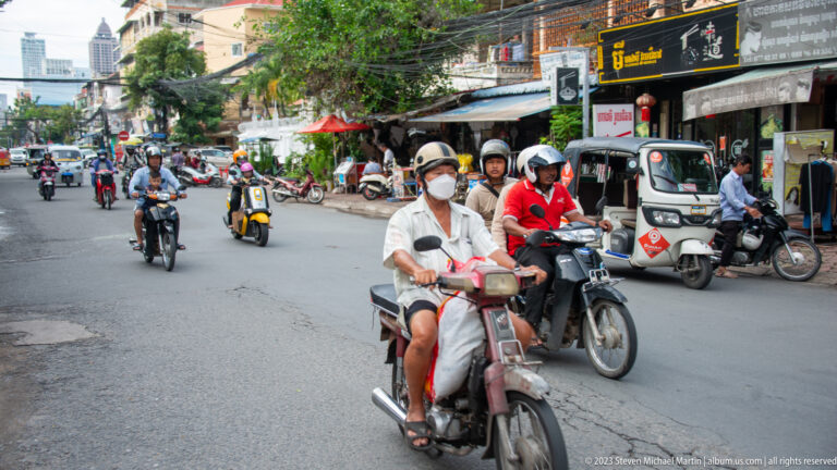 Crowded Phnom Penh street filled with motorcyclists, tuk-tuks, parked vehicles, and roadside shops.