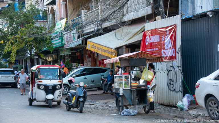 Street food cart beside a tuk-tuk, motorcycles, corrugated shopfronts, and advertising banners.