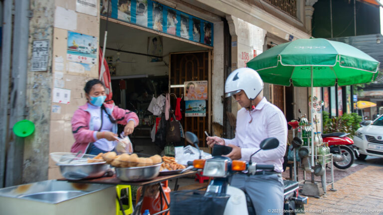 Masked vendor serves fried snacks from a curbside cart to a customer seated on a motorbike.