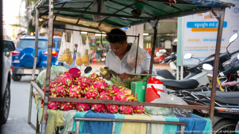 Vendor behind a pushcart piled high with bright red dragon fruit under a shaded canopy.
