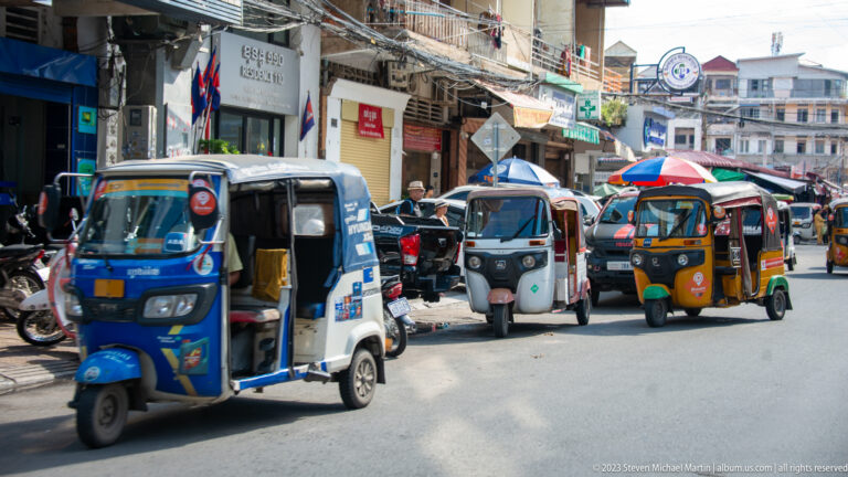 Blue, black, and orange motorized tuk-tuks traveling together along a busy commercial street.