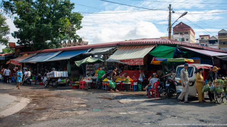Open-air neighborhood market with corrugated roofs, produce stalls, scooters, vendors, and customers at the entrance.