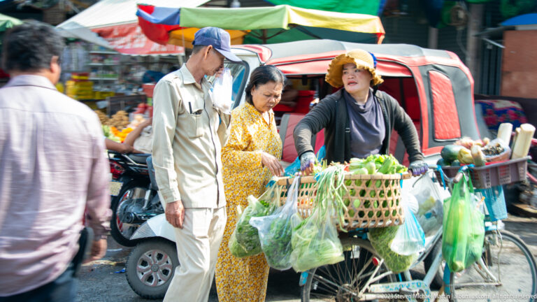 Woman pushes a bicycle loaded with baskets and bags of vegetables through a crowded market street.