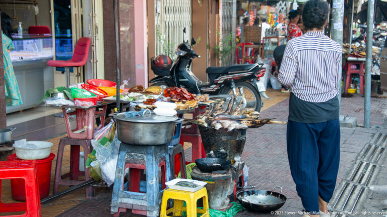Vendor works at a sidewalk food stall with charcoal braziers, metal pots, trays, and plastic stools.