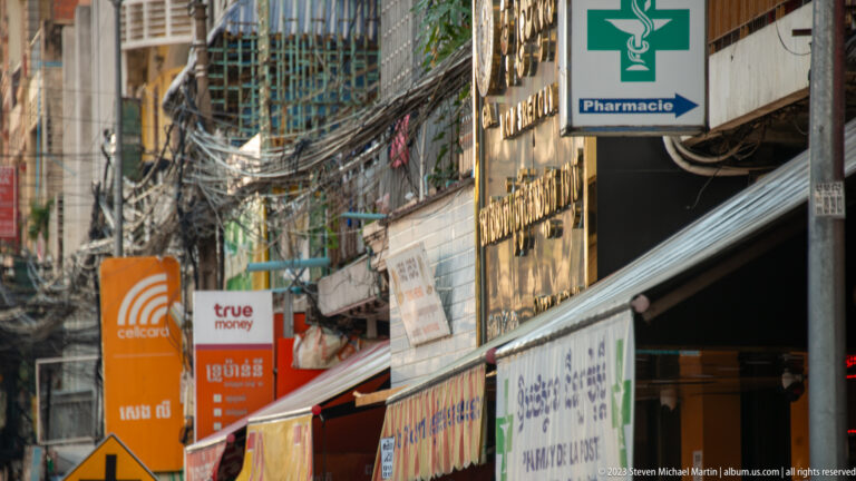 Pharmacy and mobile-payment signs amid shopfronts, metal grilles, awnings, and dense overhead wiring.