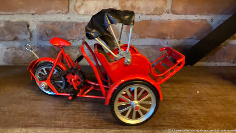 Red metal miniature three-wheeled vehicle with exposed frame and black seat on a wooden surface.
