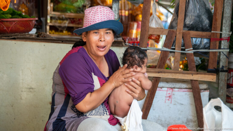 Woman in a brimmed hat cradles an infant beside a rough wooden structure at a market stall.
