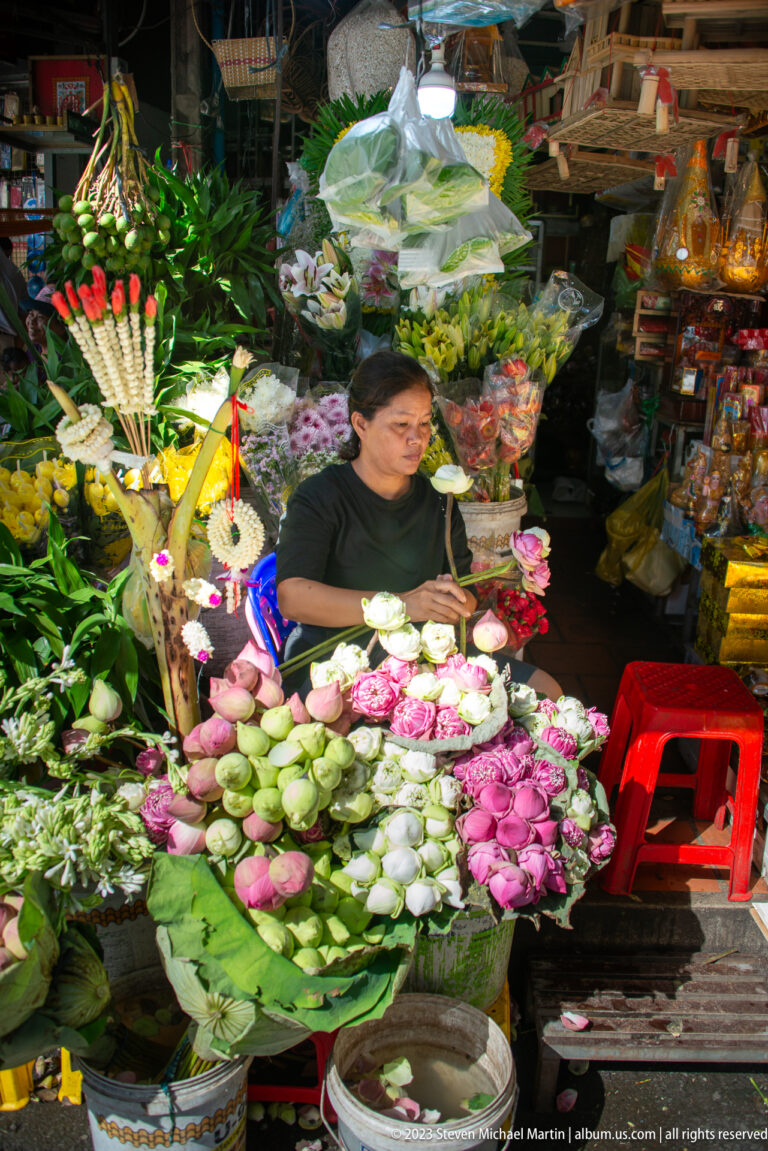 Vendor seated behind pink and white lotus blossoms, buds, folded leaves, and decorative floral offerings.