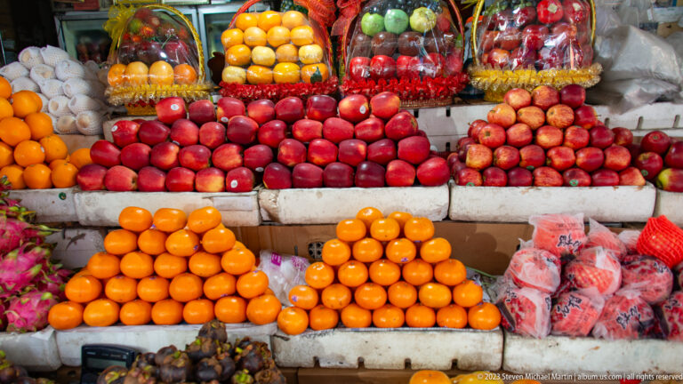 Stacks of oranges and red apples arranged in pyramids beneath hanging packages of fruit.