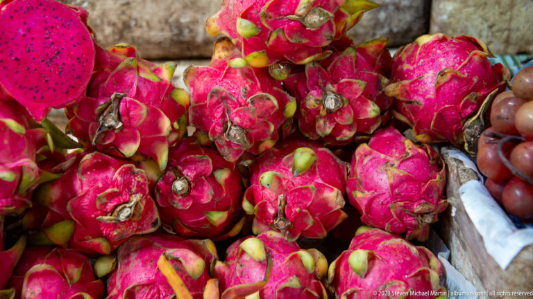 Close view of ripe pink dragon fruit with green-tipped scales piled beside other produce.