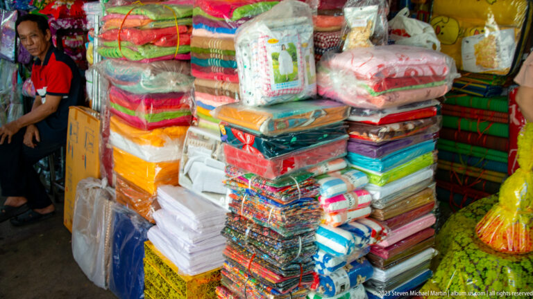Stacks of colorful folded and plastic-wrapped textiles filling a narrow indoor market stall.