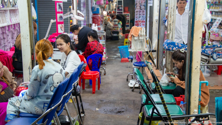 Women seated in plastic chairs beside mirrors and beauty workstations in a narrow market aisle.