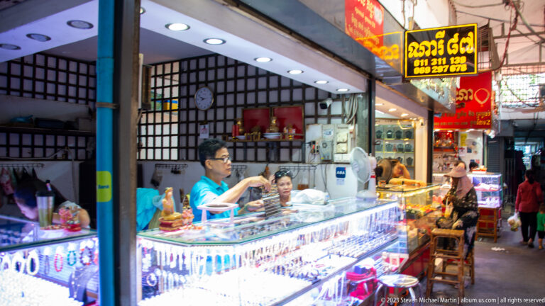 Jewelry shop with illuminated glass counters, Khmer signage, shopkeepers, and trays of ornaments.