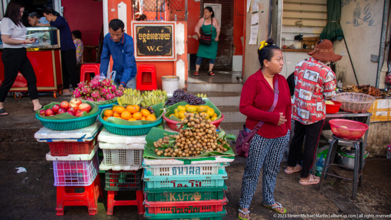 Woman standing beside a fruit stall arranged on plastic crates with oranges, longans, apples, and dragon fruit.