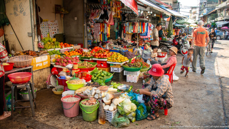 Narrow outdoor market lane crowded with seated vendors, baskets, tubs, vegetables, fruit, and hanging goods.