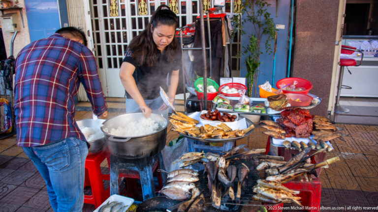 Vendor cooking over a steaming pot beside trays of grilled fish, sausages, meat, and prepared dishes.