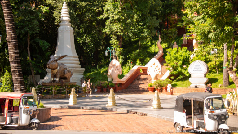 Wat Phnom entrance with animal sculpture, white stupa, ceremonial stairs, dense trees, and a parked tuk-tuk.