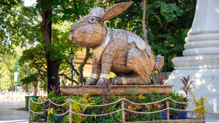 Large woven rabbit sculpture beside a white stupa base among trees and garden plants.