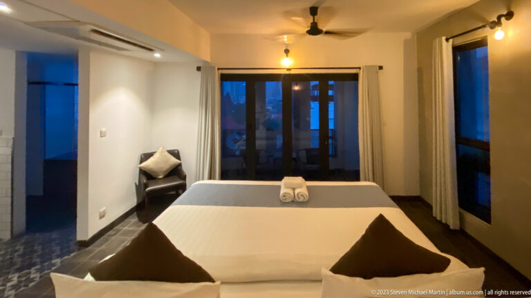 Modern hotel room with double bed, folded towels, dark pillows, ceiling fan, and glass balcony doors.