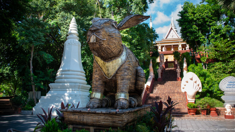 Large seated woven animal sculpture beside a white stupa and Wat Phnom staircase.