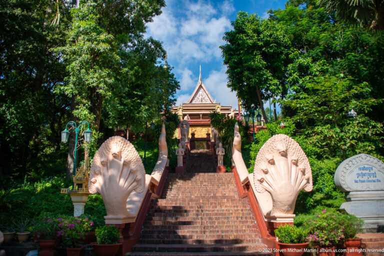 Brick staircase at Wat Phnom flanked by white multi-headed naga balustrades and dense greenery.