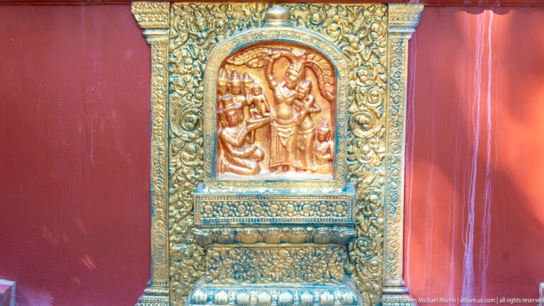 Small gilded relief of several figures set inside an ornate gold frame on a red wall.