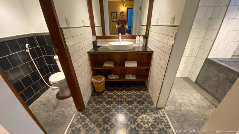Compact tiled bathroom with vanity, open shelves, toilet, glass shower, and photographer reflected in the mirror.