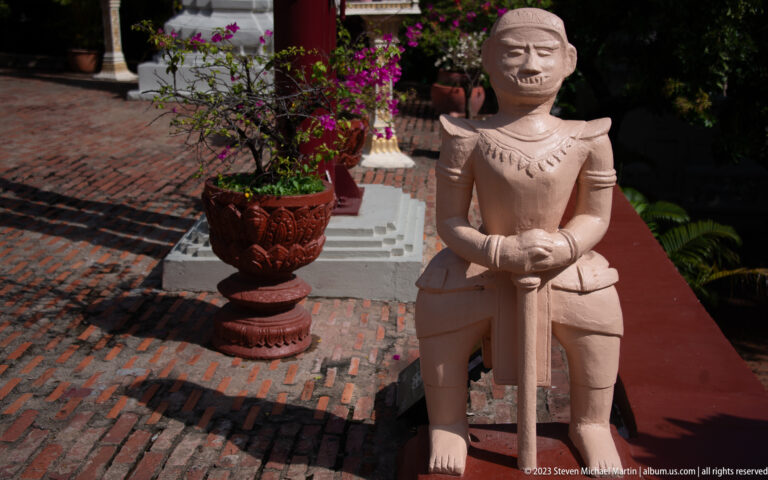 Stone-colored guardian warrior holding a sword beside a brick path and flowering plants.
