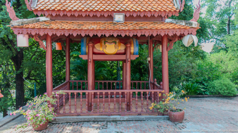 Red wooden pavilion with tiled roof sheltering a large painted ceremonial drum.