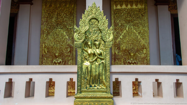 Gilded standing figure with flame-shaped halo before ornate gold relief panels on a temple façade.