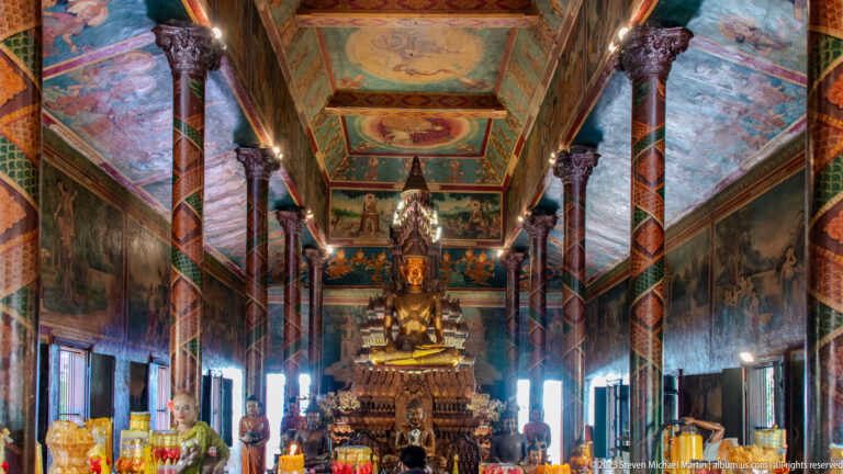 Long Wat Phnom interior with painted columns, wall murals, ceiling panels, offerings, and a central Buddha altar.