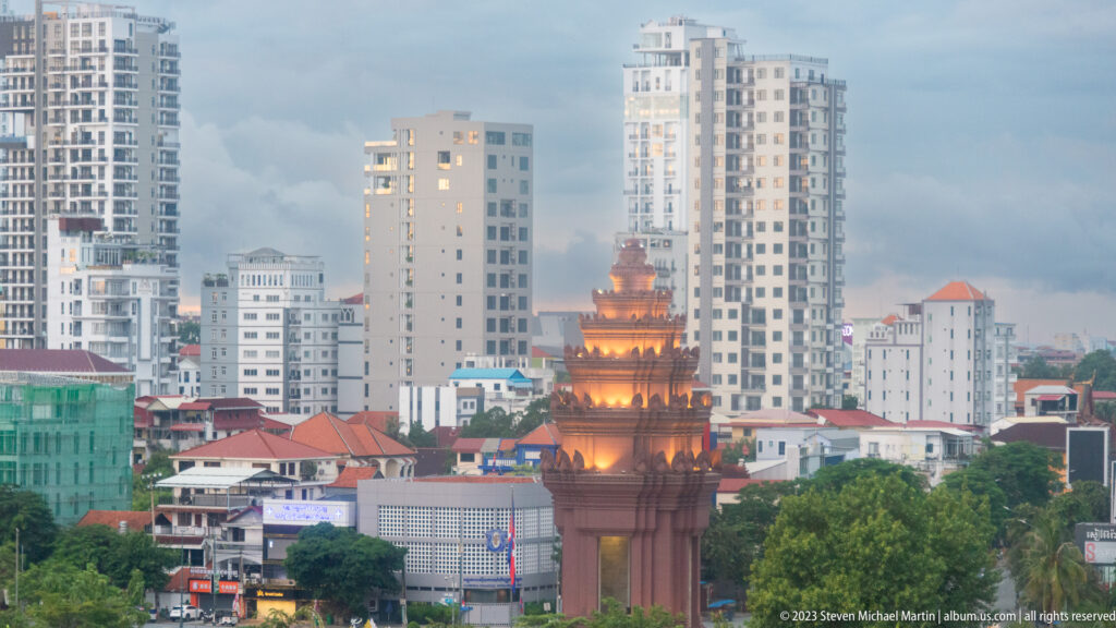 Illuminated Independence Monument framed by trees, low rooftops, and modern high-rise buildings in Phnom Penh.