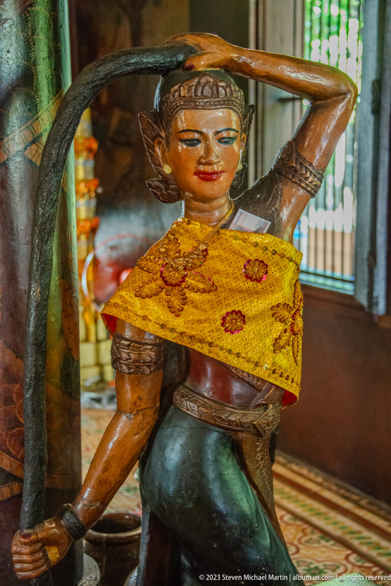 Painted standing temple figure in a yellow sash and green garment supporting an object overhead.