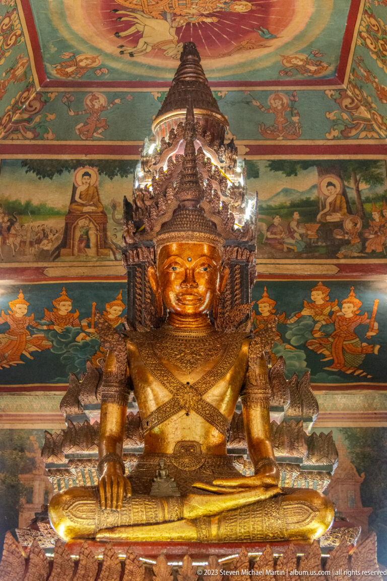 Bronze-toned crowned figure seated cross-legged before murals of worshippers and lotus motifs.