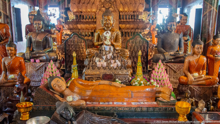 Orange reclining Buddha before seated statues, lotus offerings, carved thrones, and ritual vessels.
