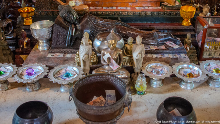Metal offering bowls, raised trays, small statues, and containers holding currency before a temple shrine.