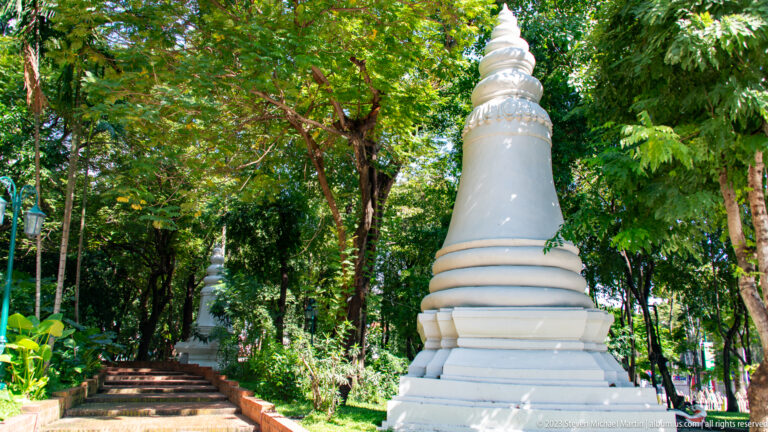 Large white stupa beside a shaded path with another stupa visible among dense trees.