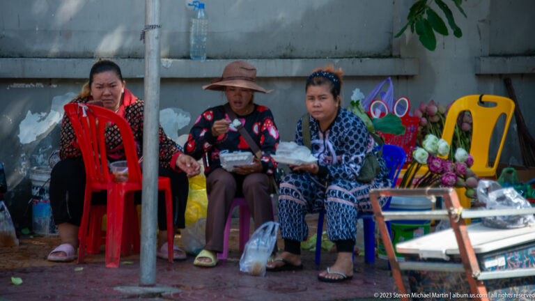 Three women seated on colorful plastic chairs beside flowers and small roadside goods.