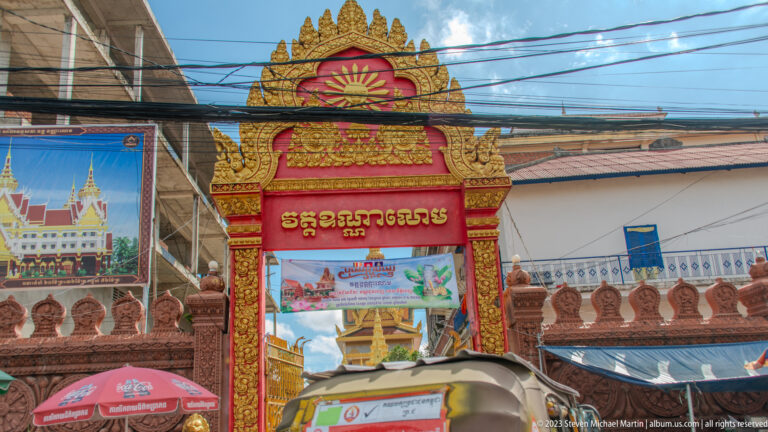 Red-and-gold Khmer temple gateway with script, lotus ornament, overhead wires, and street umbrellas.