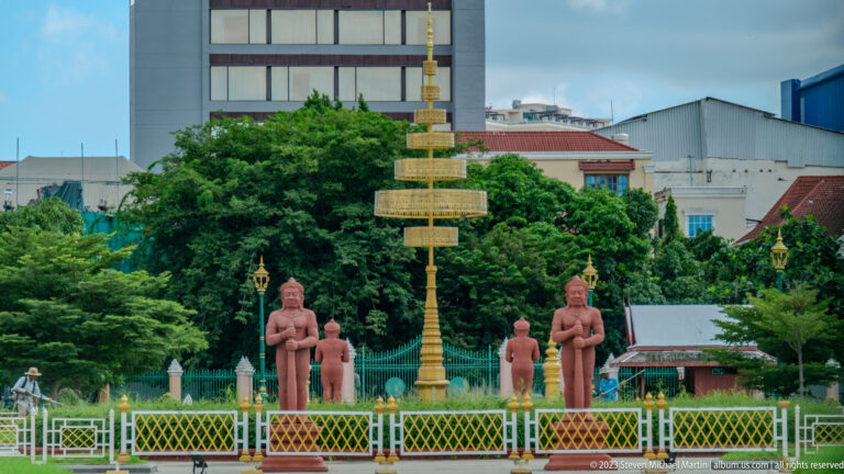 Golden ceremonial standard surrounded by reddish standing figures inside a fenced urban monument.