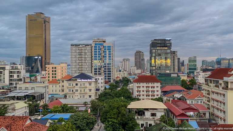 Elevated Phnom Penh skyline with modern towers, older hotels, red roofs, trees, and dark clouds.