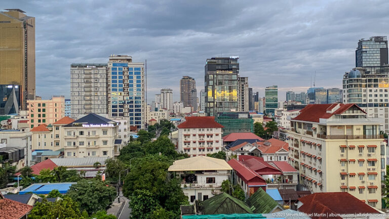 Central Phnom Penh view with tree-lined streets, red roofs, mid-rises, and a dense high-rise skyline.