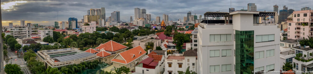 Wide Phnom Penh city view with red-roofed compounds, a white foreground building, trees, and distant towers.