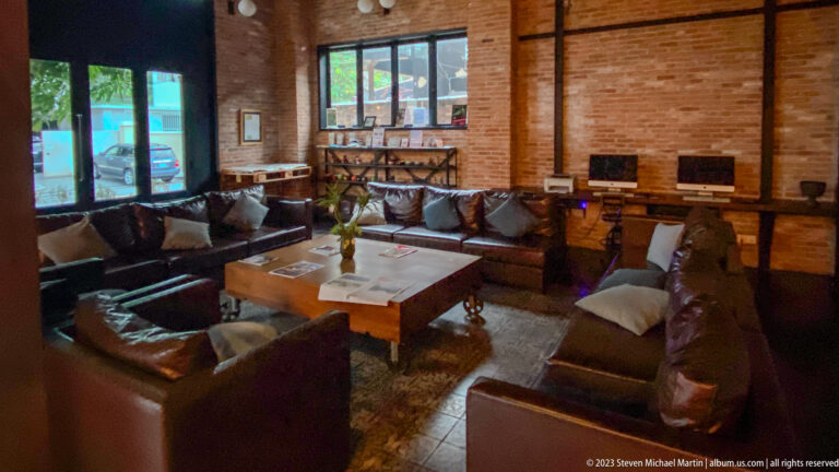 Brick-walled lounge with dark leather sofas, large wooden table, rugs, windows, and computer stations.