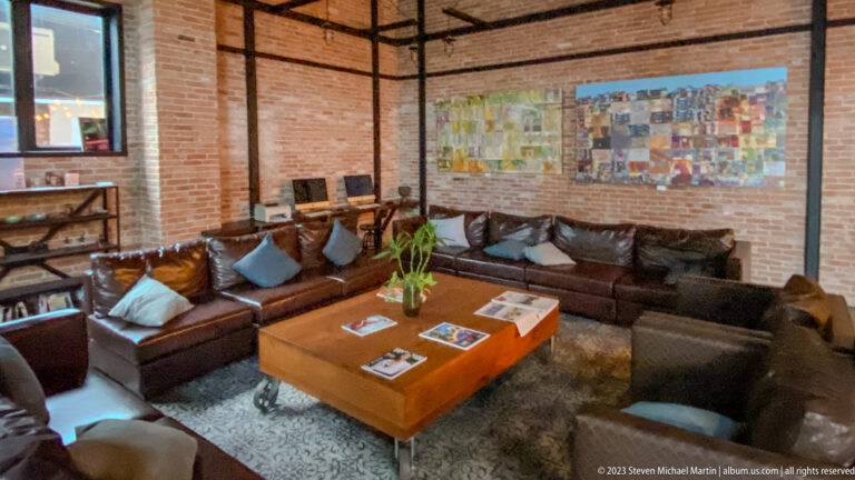 Large leather sectional around a low wooden table beneath a colorful wall collage in a brick lounge.