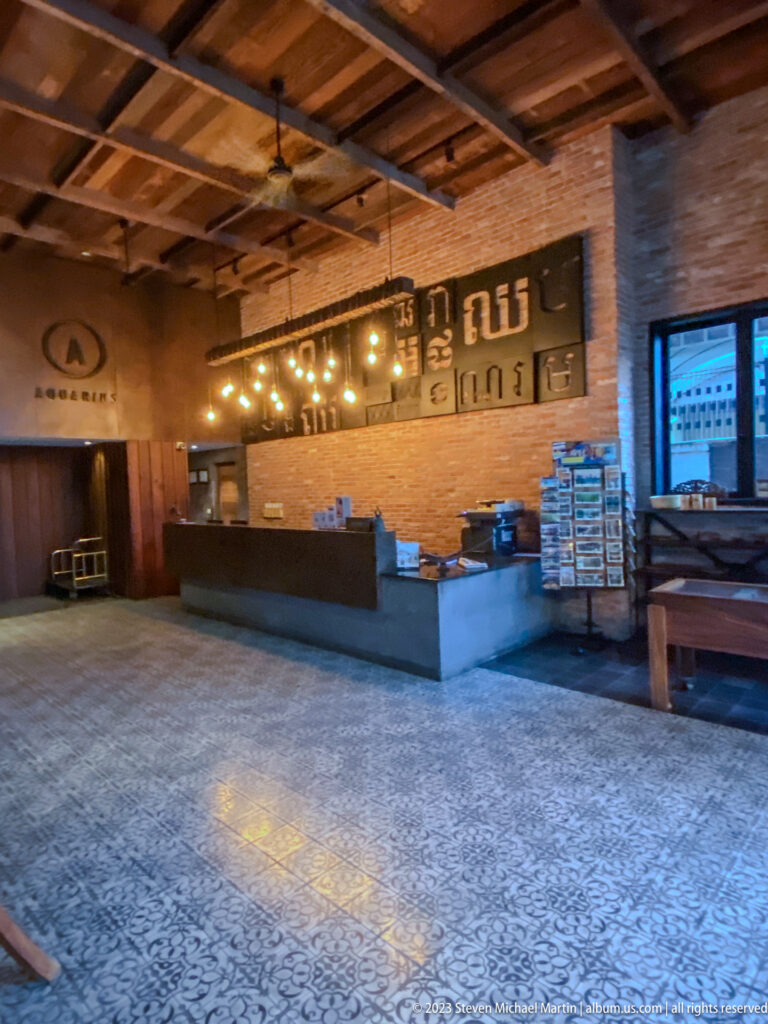 Wide brick interior with long service counter, pendant lights, Khmer signage, patterned floor, and stacked supplies.